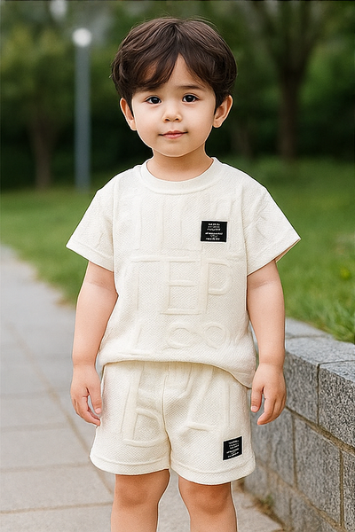 Un jeune garçon vêtu d'un ensemble d'été léger de la marque AURORE LUXURE, comprenant un haut blanc à lettres et un short. Parfait pour les enfants de 4 à 7 ans. Ensemble de vêtements d'été pour jeunes garçons.