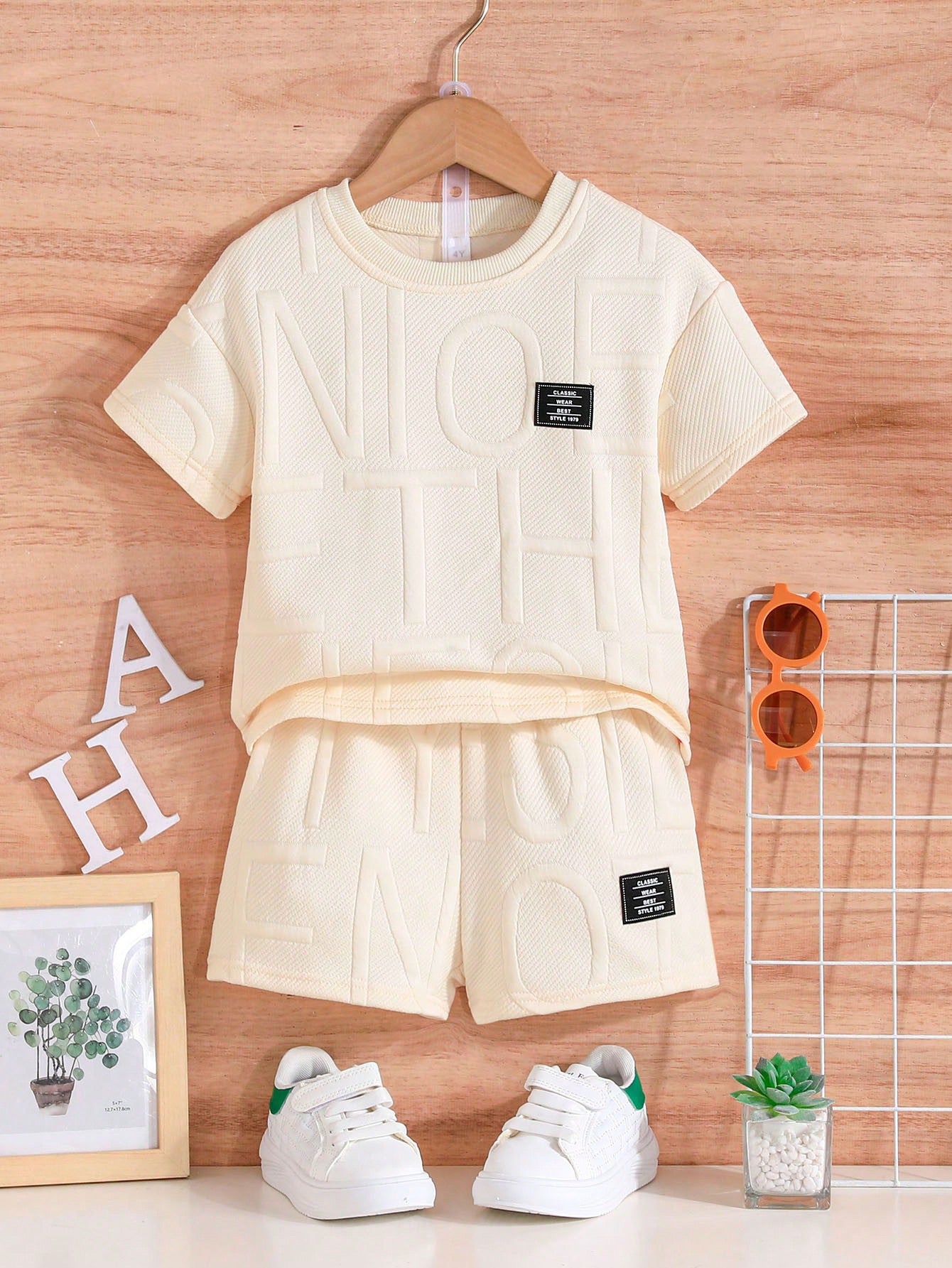 Un ensemble d'été blanc de la marque AURORE LUXURE exposé sur un cintre, comprenant un haut à lettres et un short assorti. Conçu pour les enfants de 4 à 7 ans. Ensemble d'été pour jeunes garçons.